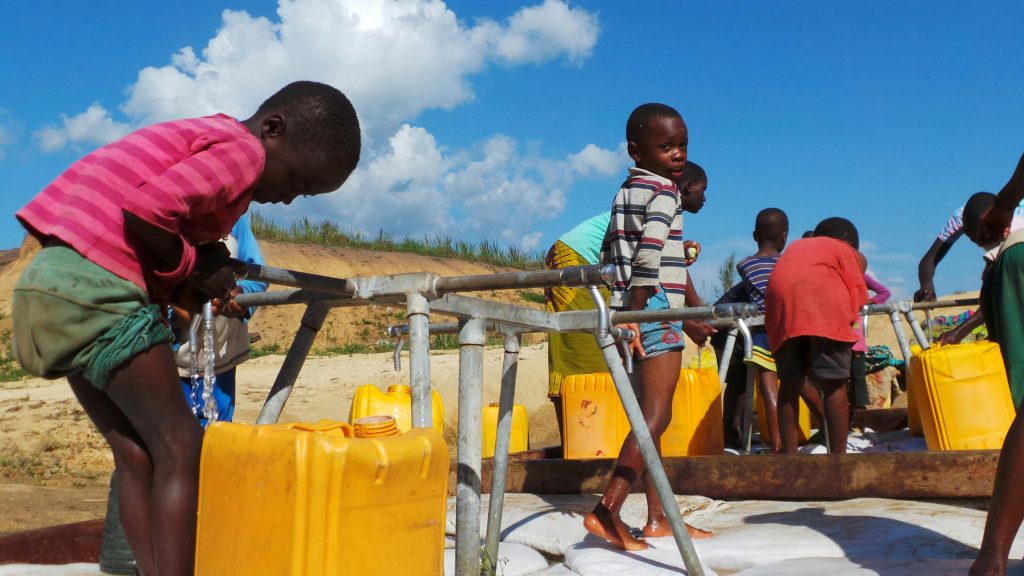Group of children filling water jugs at an outdoor faucet on a sunny day.